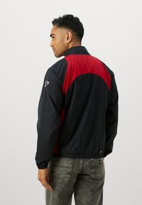 Veste coupe-vent légère, noire et rouge, avec un col haut, des poignets élastiques et un design en blocs de couleurs incurvés. Dispose de poches latérales.