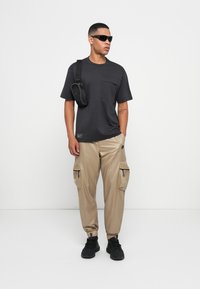Mand iført sorte solbriller, mørkegrå t-shirt, beige cargo bukser, sorte sneakers og en sort crossover taske mod en ensfarvet hvid baggrund.