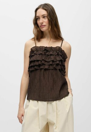 Femme aux longs cheveux bruns portant un top sans manches marron foncé à volants et un pantalon ample beige clair, debout devant un fond uni.