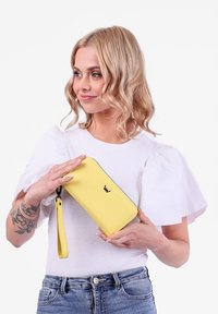 Yellow leather wallet with a sleek rectangular shape, embossed logo, and a detachable wrist strap, held against a white background.