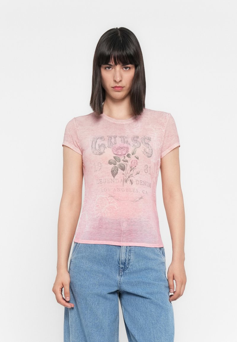 Fată tânără cu păr drept și închis la culoare, purtând un tricou grafic transparent roz cu imprimeu floral și blugi deschiși la culoare, cu talie înaltă, stând în fața unei fundaluri albe.