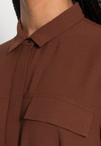 Chemise marron à col, fabriquée dans un tissu doux et texturé. Elle présente une couture verticale et une poche poitrine avec des détails de couture subtils.