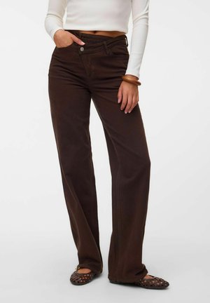 Personne portant un pantalon large taille haute marron foncé, un top court blanc, un bracelet en bois et des chaussures plates foncées perforées, debout avec une main dans la poche.