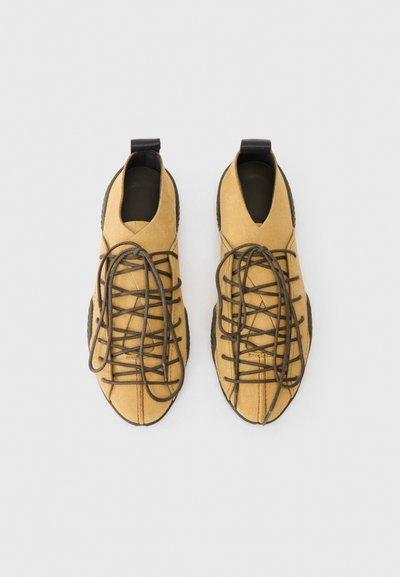 Ein Paar helle Wildleder-Schnürschuhe mit schwarzen Sohlen und Schlaufen an der Ferse, von oben auf einem weißen Hintergrund fotografiert.