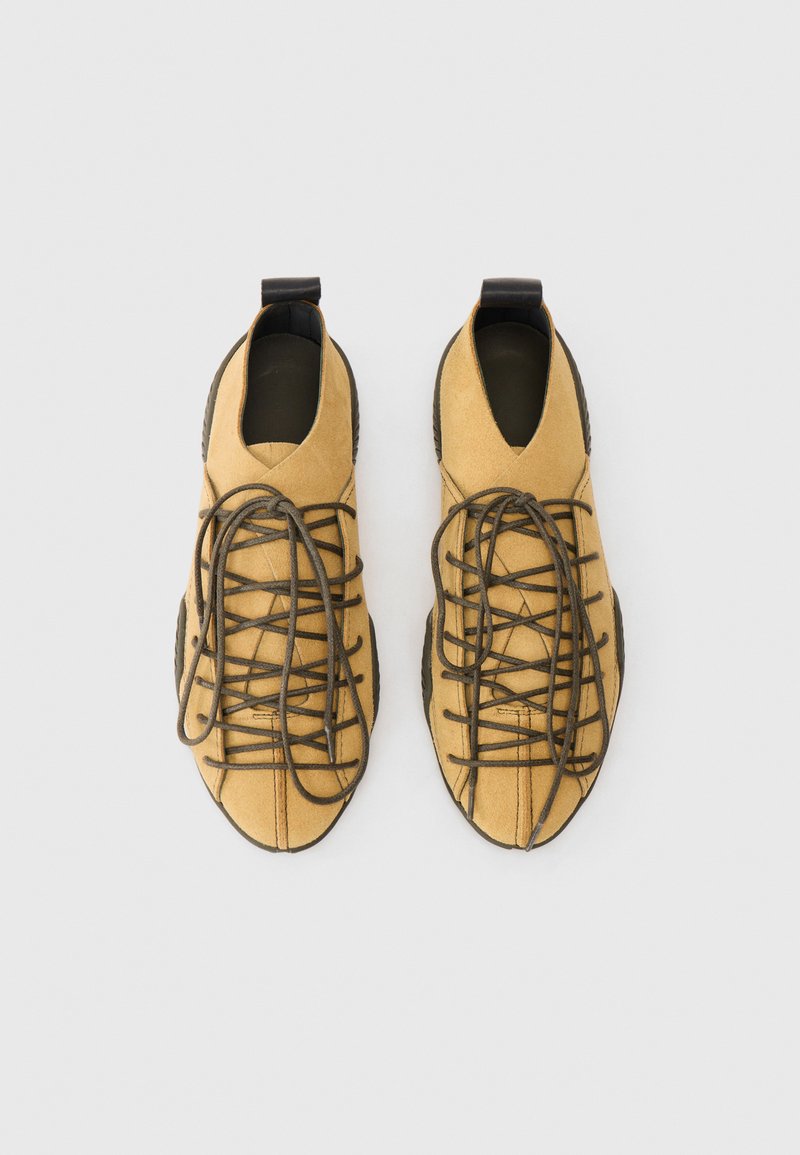 Une paire de chaussures en daim beige à lacets avec des semelles noires et des boucles au talon, photographiée de dessus sur un fond blanc.