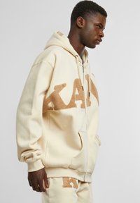 Beige hettegenser med kengurulomme; har en teksturert brun "KAL"-logo foran; ribbestrikkede ermer og hem.