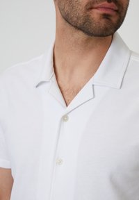 Camisa polo blanca de manga corta con cuello con muesca y dos botones. Hecha de un tejido texturizado, que presenta un sutil patrón acanalado.