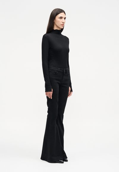 Γυναίκα με μακριά σκούρα μαλλιά, φορώντας εφαρμοστό μαύρο ζιβάγκο και μαύρο καμπάνα παντελόνι, στέκεται πλαγίως σε ένα απλό λευκό φόντο.