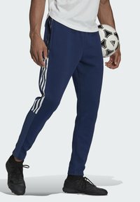 Navy athletic pants with white three-stripe accents, featuring a tapered fit and side pockets, paired with black soccer cleats and a ball.