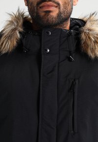 Svart parkas med en huva som är fodrad med päls, med tryckknappar och dragsko-stängningar, en ficka med dragkedja på bröstet och en strukturerad tyg.