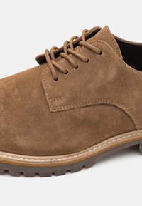 Scarpa in suede marrone con lacci, dalla consistenza liscia, dettagli cuciti, punta rotonda e suola in gomma spessa e con motivo.