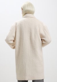 Cappotto di lana beige chiaro con una texture morbida, caratterizzato da un ampio colletto e spalle scese. Taglio rilassato, leggermente over-size.
