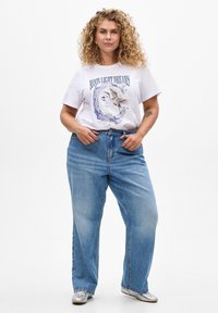 Weißes T-Shirt mit einem Grafikdesign "Moonlight Dreams", kombiniert mit hellblauen, lässig geschnittenen Jeans. Das Model trägt metallic silberne Schuhe.