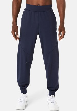 Jogging en néoprène bleu marine avec une texture lisse, une silhouette ajustée et des poches latérales, associés à des baskets blanches avec une semelle texturée.