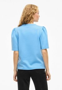 Haut bleu clair à manches courtes avec épaules bouffantes, en tissu lisse. Présente un col rond et une coupe décontractée.