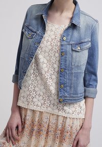 Light blue denim jacket with two front pockets and gold buttons, layered over a cream lace top and a floral-patterned skirt.