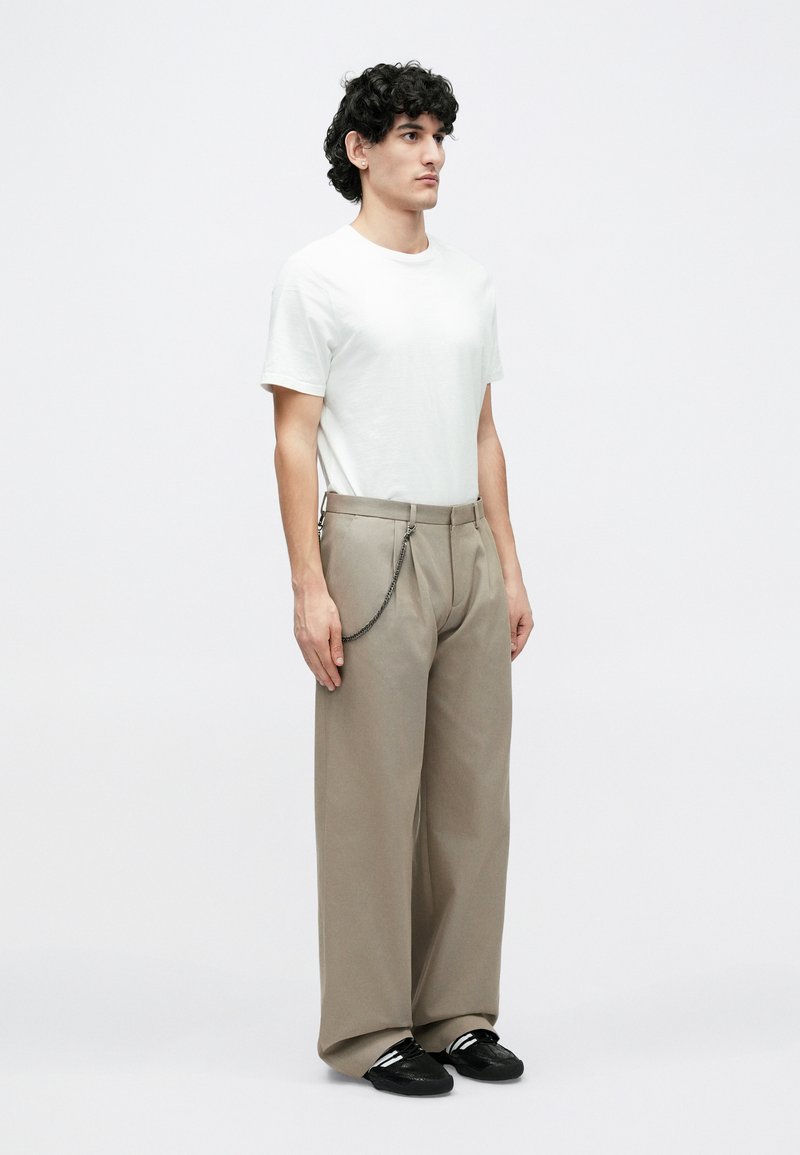 Young man with curly hair wearing a white t-shirt, beige wide-leg pants with chain accessory, and black sneakers, standing against a white background.