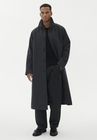 Mörkgrå överrock med hög krage, fem knappar och avslappnad passform, bärs över en svart tröja och lösa svarta byxor. Svarta skor.