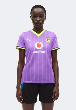 Personne portant un maillot de football violet du Borussia Dortmund avec les logos Vodafone et Puma, associé à un short noir, debout devant un fond uni.