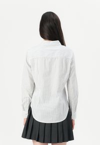 Woman with long dark hair wearing a white vertical striped shirt and a black pleated skirt, facing away from the camera against a white background.