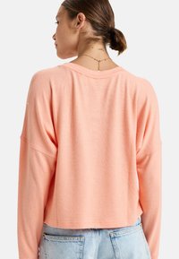 Helles korallenfarbenes, kurz geschnittenes Sweatshirt mit langen Ärmeln, weichem Textil, weitem Ausschnitt und überschnittenen Schultern, kombiniert mit hellblauen, hoch geschnittenen Jeans.