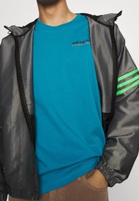 Person wearing a teal Adidas Adventure t-shirt under an unzipped shiny dark gray jacket with green stripes on the sleeve, hand in pocket.