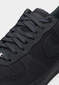 Close-up af sort Nike-sneaker med vævet tekstur, tykke snørebånd, syede overlays og præget swoosh-logo på siden.
