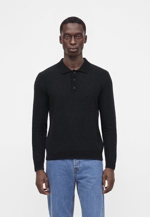 Mand iført en sort tekstureret langærmet polo og lyseblå jeans stående mod en ensfarvet hvid baggrund.