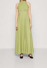 Woman wearing a floor-length, sleeveless, light green halter dress with ruffle details and black Mary Jane shoes on a plain gray background.