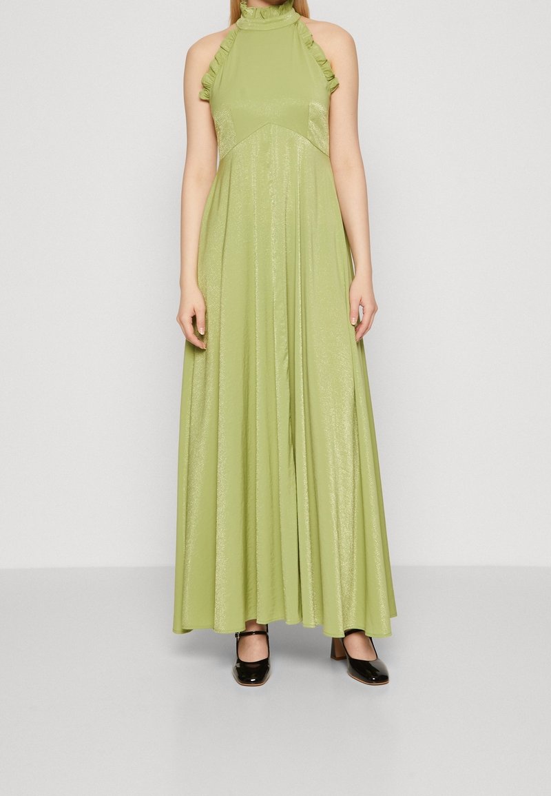 Woman wearing a floor-length, sleeveless, light green halter dress with ruffle details and black Mary Jane shoes on a plain gray background.