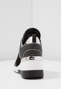 Bottines compensées noires et blanches avec accent argenté et logo Michael Kors sur le talon, présentées sur une surface blanche contre un fond uni.