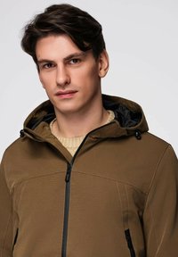 Jeune homme aux cheveux bruns portant une veste à capuche marron sur un pull en tricot beige, faisant face à l'avant avec une expression neutre.