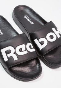 Sandales slides Reebok Classic noires avec logo blanc en gras sur des larges brides et semelles texturées sur fond blanc.