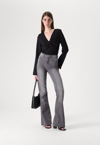 Juodas ilgomis rankovėmis viršus su surišta priekiu, derinamas su pilkomis aukštu juosmeniu platėjančiomis džinsomis ir juodomis aukštakulnėmis sandalais, laikant juodą petinę rankinę.