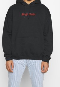 Persoon gekleed in een zwarte hoodie met "Night Addict" in rode gotische tekst, gecombineerd met blauw-witte verticaal gestreepte broek.