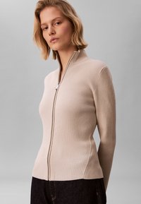 Suéter beige de punto acanalado con cierre de cremallera, cuello alto y corte ajustado, con una cremallera plateada y mangas largas.
