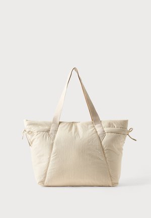 Beige blød stoftaske med brede håndtag og snøre detaljer på hver side mod en ensartet lys baggrund.