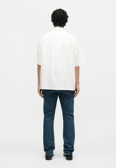 Άνδρας με σγουρά σκούρα μαλλιά, φοράει άσπρο άνετο κοντομάνικο πουκάμισο και μπλε τζιν, στέκεται γυρισμένος προς τα πίσω σε απλό φόντο.