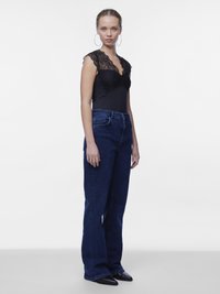 Jeune femme aux cheveux bouclés portant des boucles d'oreilles créoles, un haut en dentelle noire, un jean taille haute bleu foncé et des chaussures noires à bout pointu, debout devant un fond blanc.