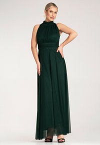 Maxi sukienka w głębokim zielonym kolorze, z wysokim dekoltem, plisowanym korpusem i zwiewnym materiałem. Przezroczysty overlay i dopasowany pas podkreślają sylwetkę.