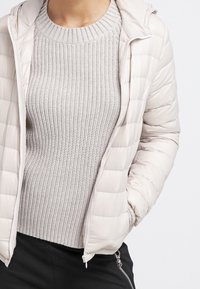 Person trägt eine hellbeige gesteppte Jacke über einem strukturierten grauen Strickpullover mit schwarzen Hosen, eine Hand in der Jackentasche.
