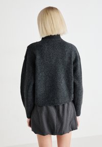 Suéter de punto gris oscuro con un cuello alto, hombros caídos y dobladillo acanalado. Presenta elementos decorativos en los hombros y una textura suave.