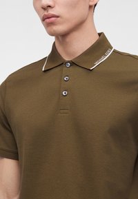 Мъж, носещ маслинено зелен поло с къс ръкав с три бутона и "Michael Kors" бродирано на яката.