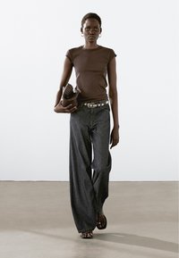 Brown fitted t-shirt, dark wide-leg denim pants, silver belt, and simple sandals. Holding a textured brown bag. Plain background.