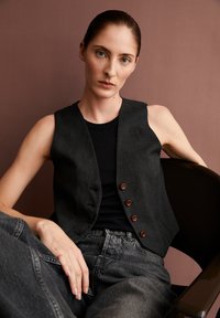 Schwarze ärmellose Weste mit fünf Holzknöpfen, getragen über einem schwarzen Rippenoberteil und kombiniert mit grauen hochgeschnittenen Jeans, sitzt auf einem Stuhl.