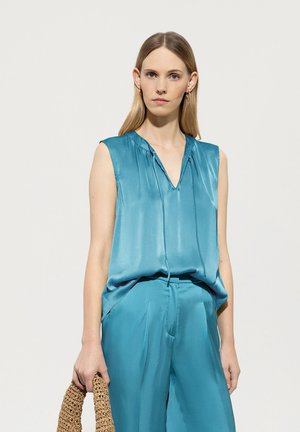 Türkises ärmelloses Top aus Satin mit V-Ausschnitt und gefaltetem Frontdetail, kombiniert mit passenden Hosen und gehalten mit einer geflochtenen Tasche.