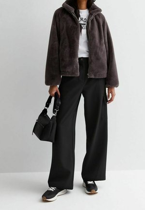 Henkilö pukeutunut tummanruskeaan vetskarilliseen fleecetakkiin, valkoiseen graafiseen t-paitaan, mustiin leveälahkeisiin housuihin, mustiin tennareihin ja pitää mustaa käsilaukkua.