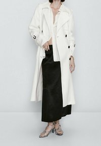 Lang hvid trenchcoat med sorte knapper, lagd over en sort satin skirt. Har en struktureret krave og brede ærmer. Sølv stilethæle.