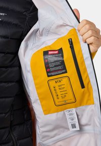 Giacca a vento nera con interno giallo. Presenta una tasca con zip, etichetta della tecnologia RECCO e istruzioni per la cura su un'etichetta bianca.