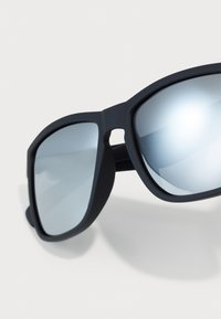 Ochelari de soare negri cu lentile supradimensionate, pătrate, având un finisaj mirror. Rama are o textură mată și un design geometric îndrăzneț.